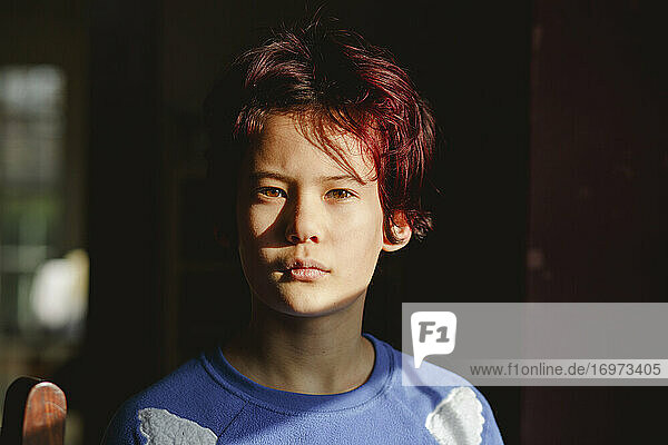 A boy with dyed red hair stands in dark room in bright patch of light