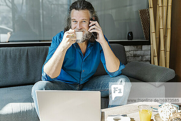 Älterer Mann mit Bart telefoniert und trinkt Kaffee sitzend