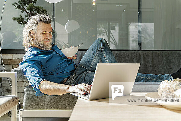 Älterer Mann mit Bart lächelt und arbeitet mit seinem Laptop auf einem Sofa