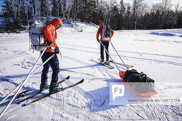 Two men attach pulk sleds to their backpacks in a snowy parking lot