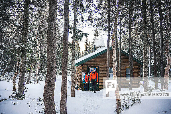 Zwei Männer mit Jacken und Rucksäcken verlassen eine Hütte in einem verschneiten Wald