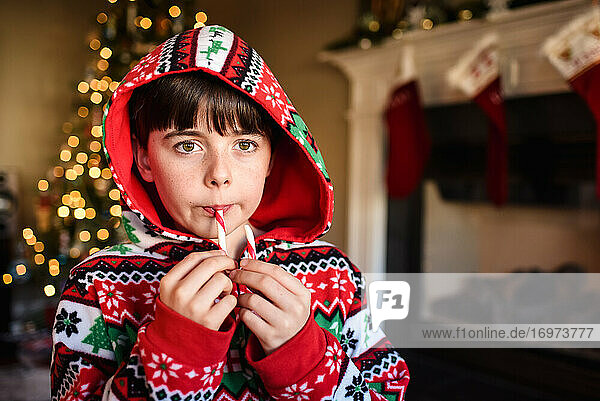Cute boy in festive pajamas eating a candy cane at Christmas time.