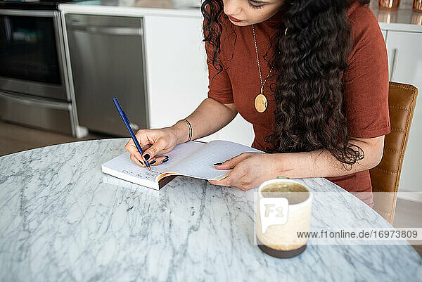 Frau mit langen Haaren schreibt am Küchentisch in ein Notizbuch