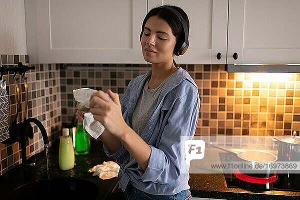 Young housewife with headphones wiping dishes in kitchen