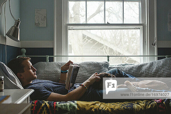 A young man lays on his bed with dog on lap reading by window light