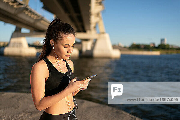 Frau mit Kopfhörern  die beim Training an der Strandpromenade auf ihrem Smartphone surft