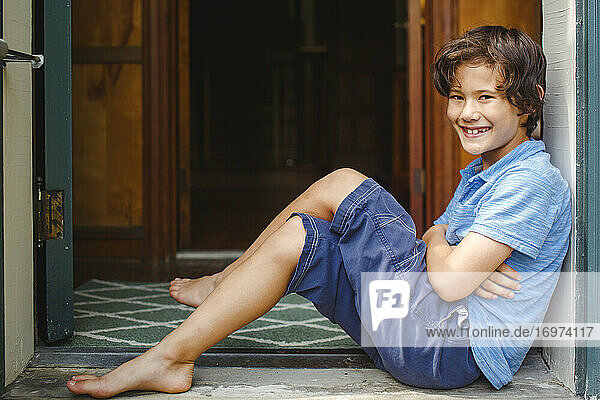 A boy with missing teeth sits smiling in a font entryway barefoot