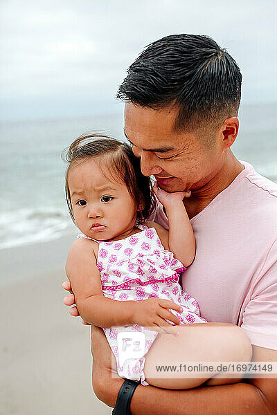 Liebevoller Vater hält süße asiatische Tochter mit pummeligen Beinen am Meer.