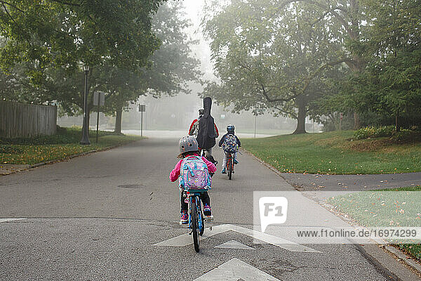A father with a cello on his back bikes in fog on street with two kids