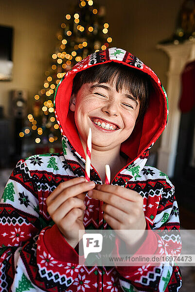 Fröhlicher Junge im festlichen Pyjama  der zur Weihnachtszeit eine Zuckerstange isst.