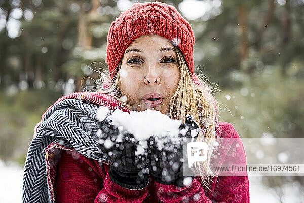 Porträt einer glücklichen jungen Frau  die im Winter mit Schnee spielt und Schneeflocken in die Kamera bläst  Kopierraum