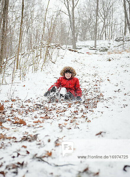 Happy boy sledding down a hill in the woods on a snowy winter day.
