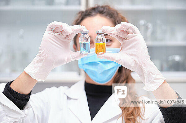 Junge Wissenschaftlerin mit Gesichtsmaske  die zwei blaue und orangefarbene Fläschchen mit Coronavirus-Impfstoff hält. Entwicklung des Impfstoffs Covid-19.