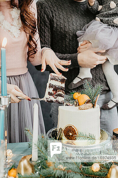 Junge Familie mit einer Tochter in festlicher Kleidung an einem gedeckten Tisch mit Kerzen  Girlanden  Wunderkerzen und einem Kuchen in der Nähe des Weihnachtsbaums an Silvester