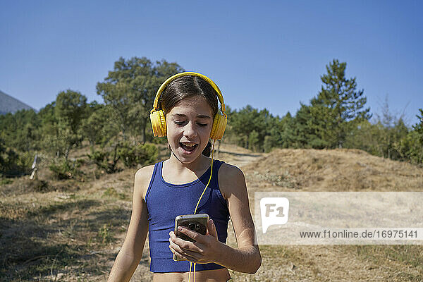 Schönes Mädchen  das mit gelben Helmen Musik hört und ein mobiles Gerät auf einem Feldweg hält