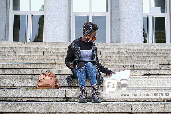 Afrikanische Studentin an der Universität  die einen Gesichtsschutz trägt und auf einer Treppe im Freien auf dem Campus sitzt. Neue Normalität im College.