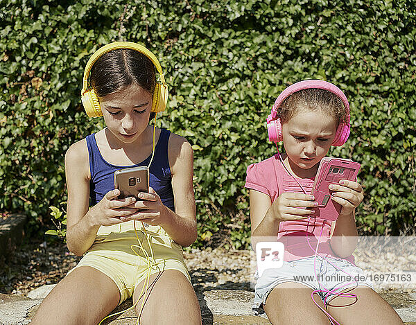 two girls looking at their smart phones and listening to music with headphones sitting on a stone wall in sunny day. Technology concept