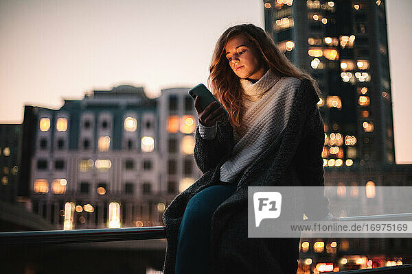 Teenage girl using smart phone sitting on railing in city at night
