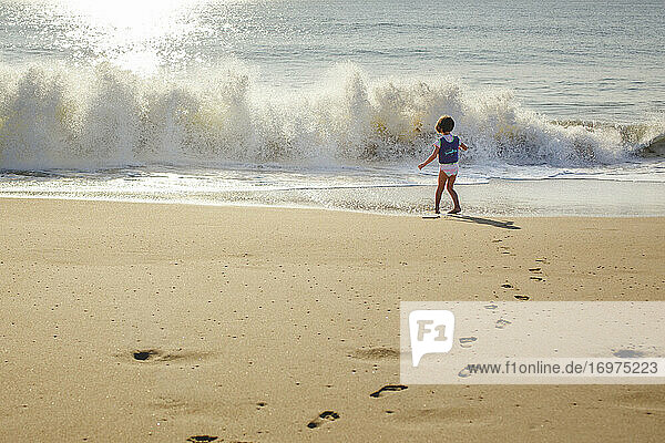 A little girl in life vest stands in front of oncoming wave at beach