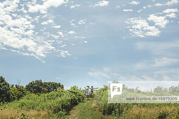 Zwei glückliche Kinder stehen mit ausgebreiteten Armen auf einem Hügel unter einem blauen Himmel