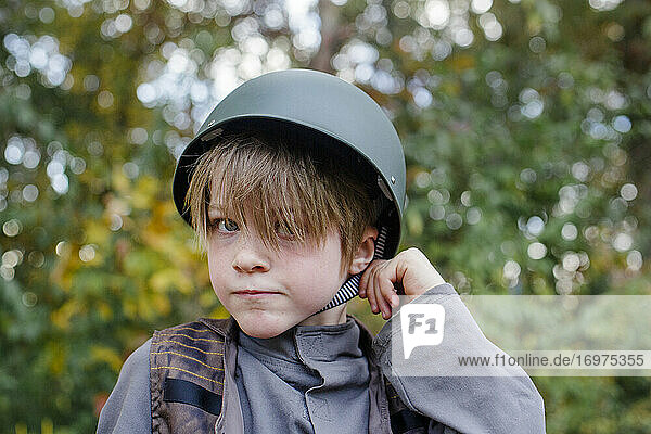 A boy dressed in a soldier costume tugs at the strap of his helmet