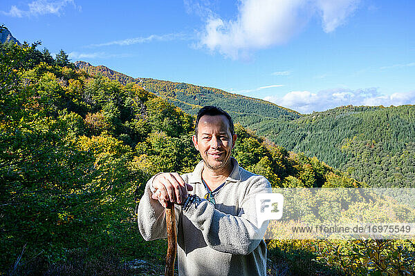 Smiling hiker man holding a pole into forest while looking at camera