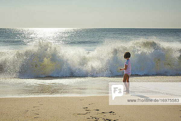 A small girl stands at edge of ocean watching wave break onto beach