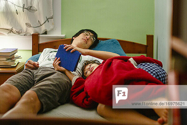 Ein Junge kuschelt sich an seinen schlafenden Vater  der zur Schlafenszeit ein Buch liest