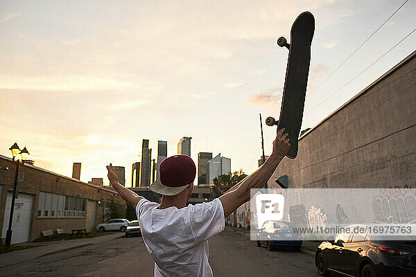 Junger Skateboarder hält sein Brett in die Luft und schaut in Richtung Innenstadt