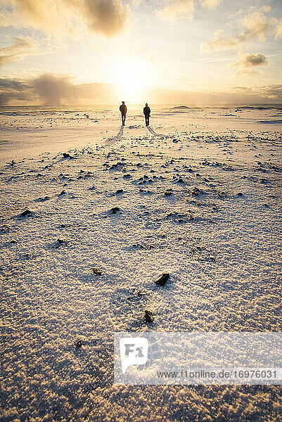 Zwei Menschen spazieren bei Sonnenaufgang am schwarzen Sandstrand in Island im Winter