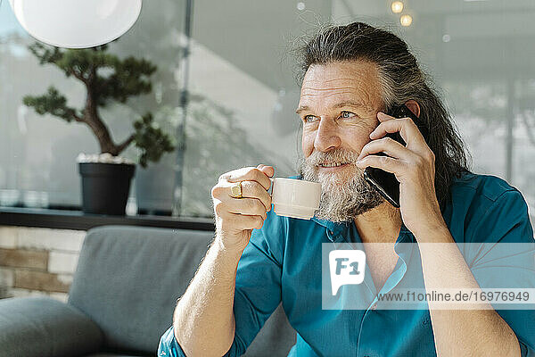 Älterer Mann mit Bart telefoniert und trinkt Kaffee sitzend