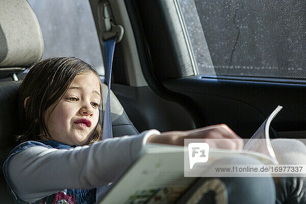 A cute little girl sits in a carseat in sunlight studying a book