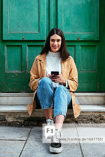 Porträt einer jungen stilvollen Frau  die im Freien sitzt und ein Smartphone benutzt