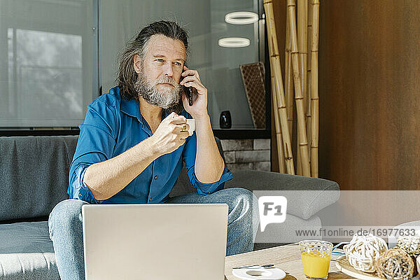 Älterer Mann trinkt einen Kaffee und telefoniert auf einem Sofa sitzend