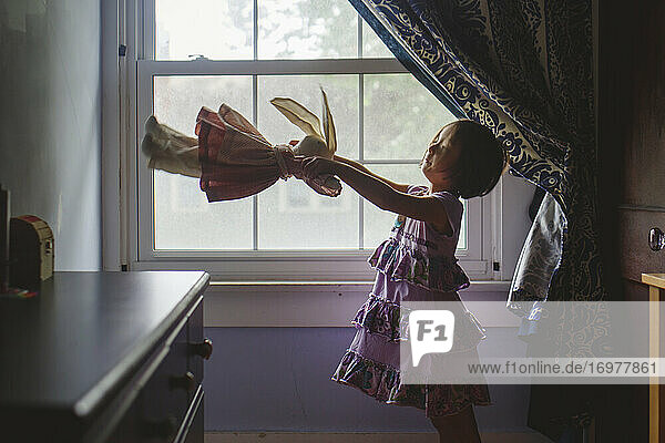 Ein süßes kleines Mädchen steht am Fenster und spielt mit einem weichen Stofftier