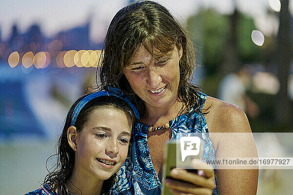 Porträt von Mutter und Tochter im Teenageralter in blauen Kleidern und Blick auf das Handy in der Nacht mit einem Strand im Hintergrund. Urlaub Konzept