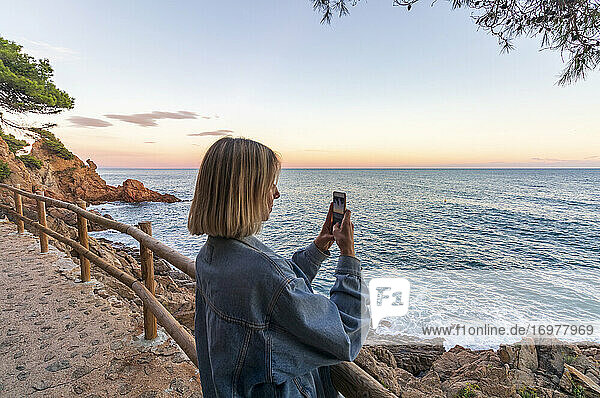 Young blonde girl photographing the sunset on the coast