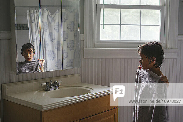 Ein Junge steht in ein Handtuch gewickelt vor der Dusche und schaut in einen Spiegel.