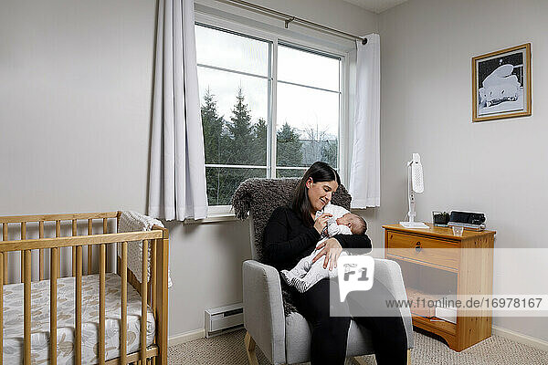 Eine Mutter sitzt in einem Schaukelstuhl in einem Kinderzimmer und füttert ihren Säugling mit einer Flasche
