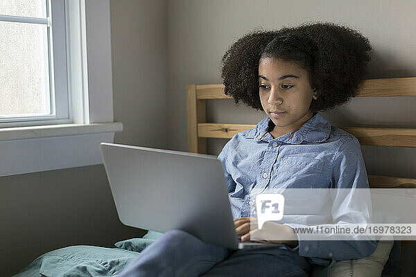 Ten year-old bi-racial girl works on her laptop sitting on bed