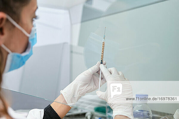 Junge Wissenschaftlerin mit Gesichtsmaske  die einen Coronavirus-Impfstoff in einer Spritze und Nadel in einer Laminar-Flow-Haube hält. Entwicklung eines Covid-19-Impfstoffs.