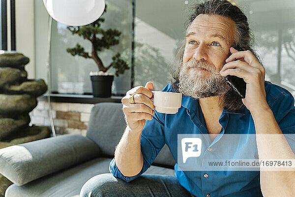 Älterer Mann trinkt einen Kaffee und telefoniert auf einem Sofa sitzend