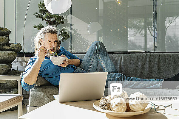 Älterer Mann mit Bart  der auf einem Sofa einen Salat isst und auf seinen Laptop schaut. Pause Zeit Konzept