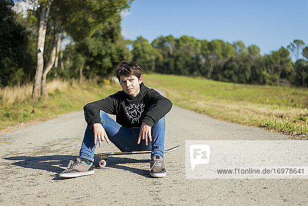 Gutaussehender Teenager mit einem Skateboard auf einer asphaltierten Straße sitzend