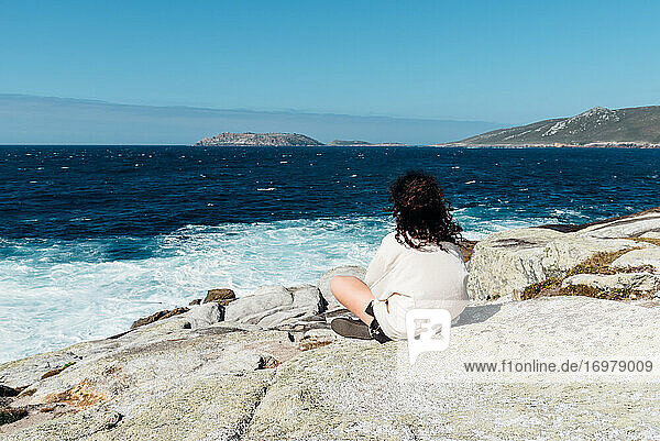 Junge Frau mit lockigem Haar sitzt auf den Felsen und schaut aufs Meer