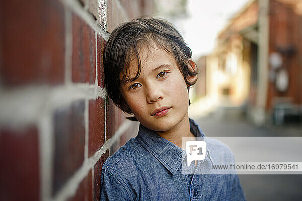 a beautiful serious boy leans against wall in sunlit brick alley