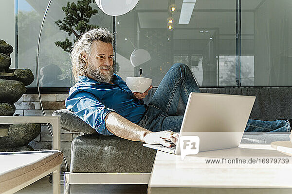 Älterer Mann mit Bart lächelt und arbeitet mit seinem Laptop auf einem Sofa