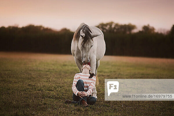 Teenager-Mädchen sitzt vor einem Pferd und schaut liebevoll zu ihm auf