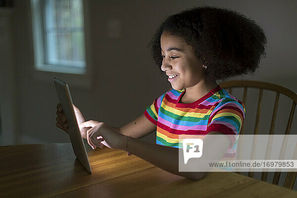 Lächelndes zehnjähriges gemischtrassiges Mädchen arbeitet am iPad am Tisch