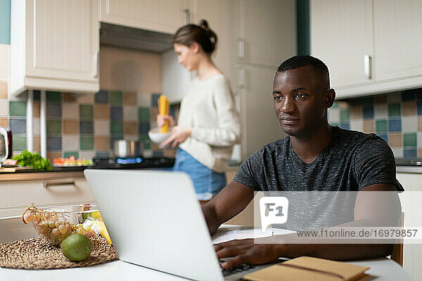 Intelligenter schwarzer Mann arbeitet am Laptop in der Nähe der kochenden Freundin